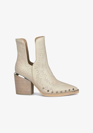Beżowe botki za kostkę z ostrym noskiem, perforowanym wzorem i srebrnymi akcentami. Posiadają drewnianą, warstwową podeszwę dla większej stabilności.
