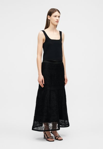 Jeune femme aux longs cheveux bruns portant un haut noir sans manches, une jupe longue en dentelle noire détaillée et des sandales noires à talons.