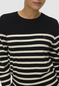 Maglione a righe nere e crema, design a girocollo, maniche lunghe, con una piccola accentuazione a forma di cuore rosso sul petto. Tessuto morbido.