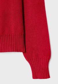 Manche et ourlet de pull rouge tricoté avec poignets côtelés sur fond gris clair, montrant la texture et les détails du tissu.
