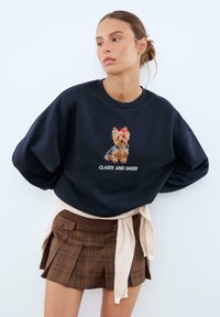 Felpa blu navy con un design ricamato di un cane e il testo "CLASSY AND SASSY". Abbinata a una gonna a quadri marrone e un maglione beige annodato.