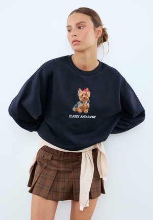 Felpa blu navy con un design ricamato di un cane e il testo "CLASSY AND SASSY". Abbinata a una gonna a quadri marrone e un maglione beige annodato.