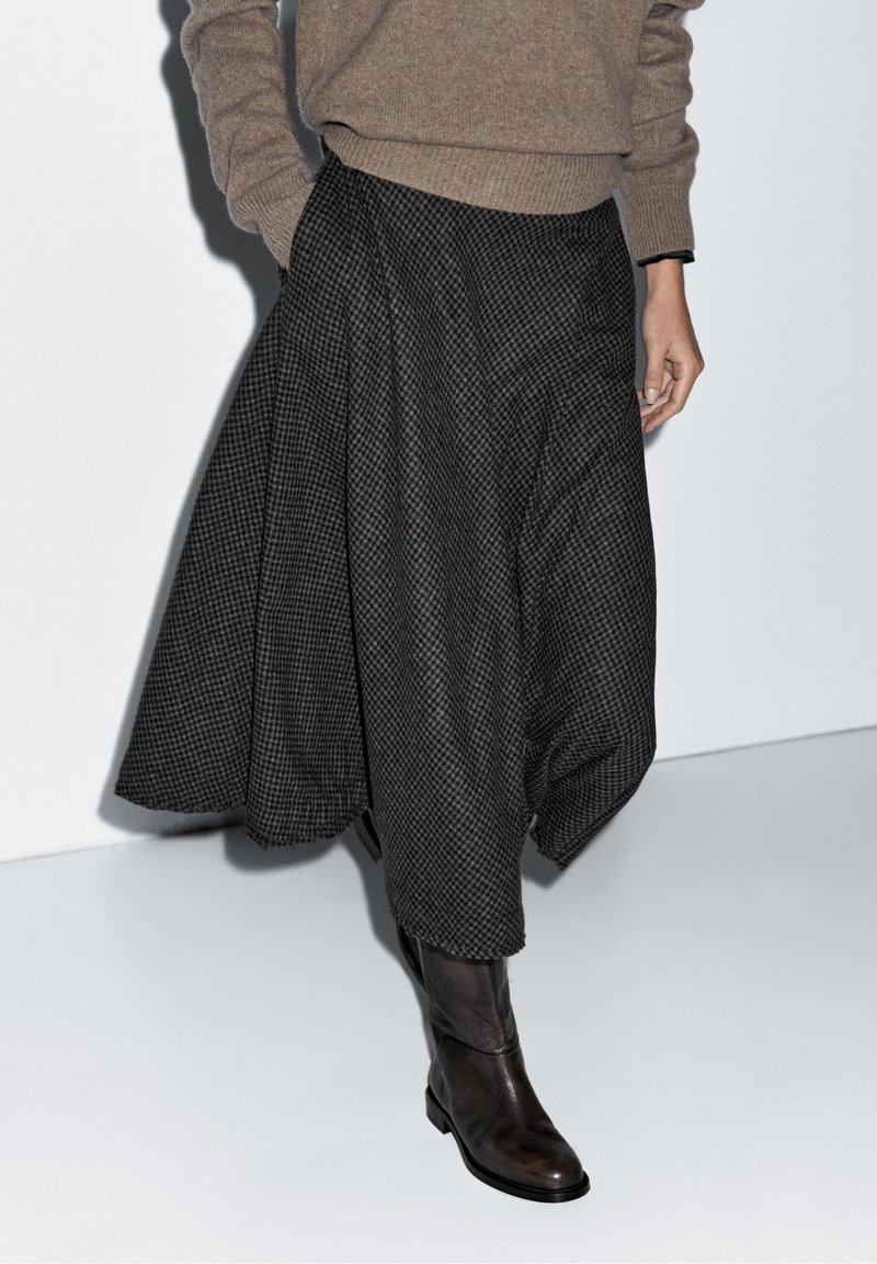 Personne portant un pull marron, une jupe midi à carreaux noirs et gris, et des bottes en cuir marron foncé, debout devant un fond clair uni.