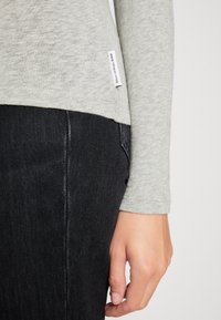 Grijze lange mouwen top met een gestructureerde afwerking, gecombineerd met donkere spijkerbroeken. Zichtbaar label op de zijnnaad. Close-up van een hand die op de taille rust.