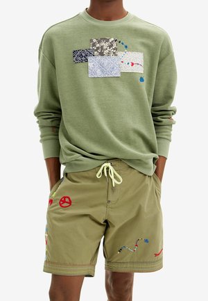 Sweatshirt vert clair avec un design en patchwork comprenant diverses textures et couleurs. Shorts olive avec des accents brodés colorés et cordon de serrage.