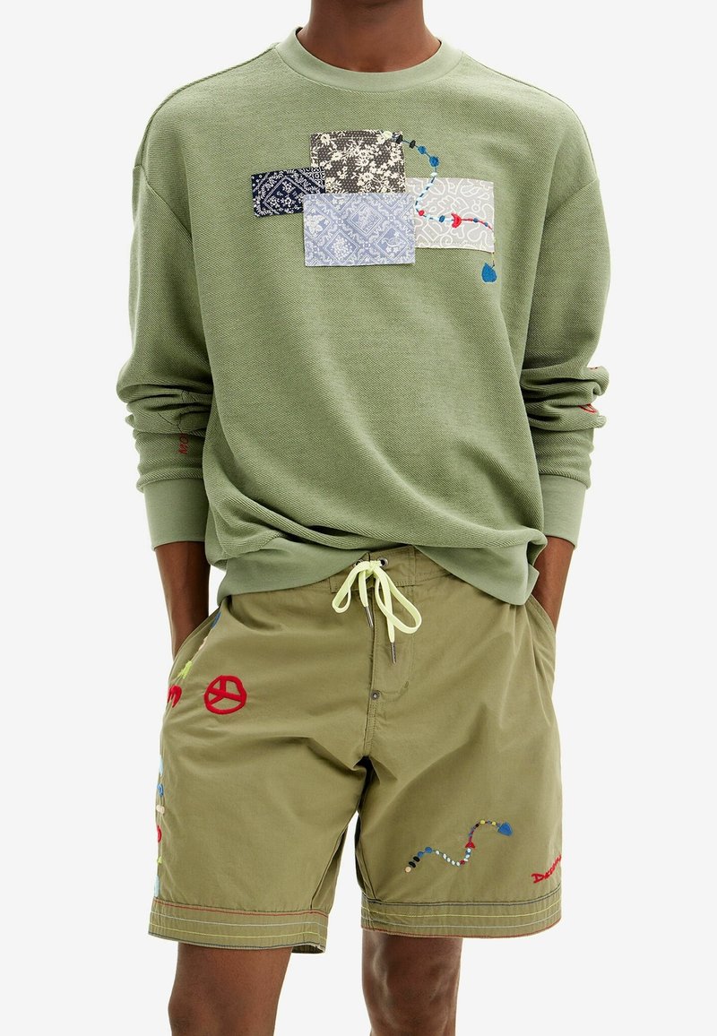 Felpa verde chiaro con design a patchwork, realizzata con diverse texture e colori. Pantaloni corti oliva con accenti ricamati colorati e coulisse.