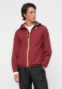 Chaqueta de chubasquero roja con cremallera al frente, presentada con llamativas rayas amarillas y blancas, capucha ajustable y puños elásticos. Pantalones negros con logo.