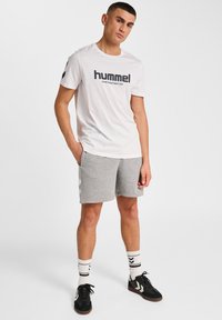 Vit Hummel t-shirt med svart logotyp, matchad med grå shorts. Vita och svarta randiga strumpor, svarta sneakers med vita detaljer.