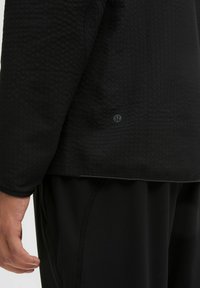 Černá texturovaná sportovní bunda s zipem na spodním lemu a logem. Materiál je hladký, s strukturovaným vzorem na povrchu.