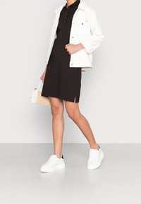 Vestito polo nero con spacchi laterali, abbinato a una giacca di jeans bianca e sneakers bianche con dettagli neri, mentre tiene una piccola borsa intrecciata.