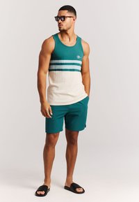 Camiseta sin mangas color verde azulado con rayas horizontales en tonos variados, mitad inferior beige; pantalones cortos a juego color verde azulado; sandalias negras deslizantes; gafas de sol marrones.