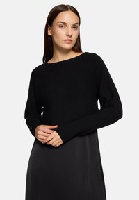 Femme aux cheveux bruns raides portant un pull noir côtelé à manches longues sur une jupe noire lisse, debout devant un fond clair uni.