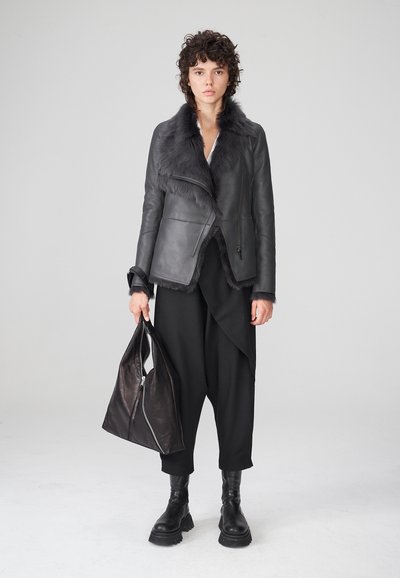 Chaqueta de cuero negra con cuello de piel sintética, dobladillo asimétrico y cierre de cremallera. La modelo sostiene un bolso negro texturizado, vistiendo pantalones negros holgados y botas.