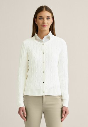 Kvinde står oprejst iført en hvid kabelstrikket cardigan med knapper over en skjorte med krave og beige bukser, vendt mod kameraet foran en ensfarvet baggrund.