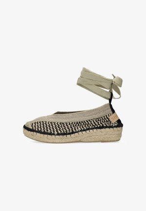 Beige en zwarte geweven espadrille met een zool van jute en een beige enkelband die aan de achterkant in een strik is gebonden.