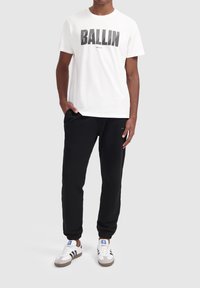 Witte katoenen t-shirt met de tekst "BALLIN" in het zwart, gecombineerd met zwarte joggingsbroeken en witte sneakers met zwarte strepen en blauwe accenten.