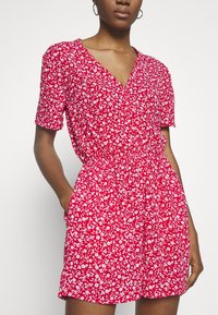 Femme portant une combinaison-short rouge à manches courtes avec un motif floral blanc, un col en V et une taille élastique, debout les mains dans les poches.
