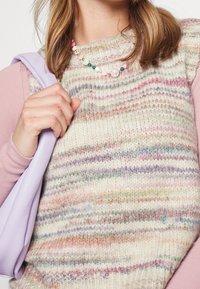 Maglione lavorato a maglia con righe multicolori in toni pastello, indossato sopra una camicia a maniche lunghe. Tieni in mano una borsa lavanda, decorata con una collana di perline.