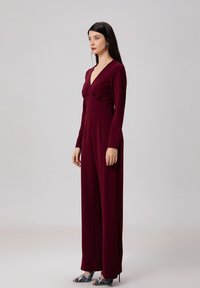 Combinaison bordeaux à manches longues et décolleté en V, avec un buste ajusté et un pantalon large et fluide. Des motifs sur les talons ajoutent de l'attrait visuel.