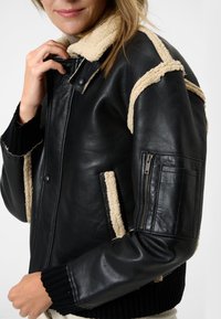Zwarte leren jas met een beige schapenvachtkraag en accenten, voorzien van geribbelde manchetten en een ritszak op de mouw.