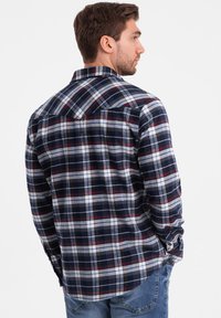 Camicia a flanella a quadri blu navy e bianca con accenti rossi, maniche lunghe, polsini con bottoni e colletto a punta, indossata con jeans di denim.