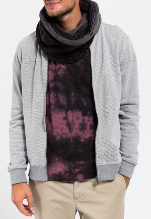 Homme portant un pantalon beige, une chemise tie-dye noire et violette, une veste zippée texturée gris clair, et une écharpe infinie en maille noire.