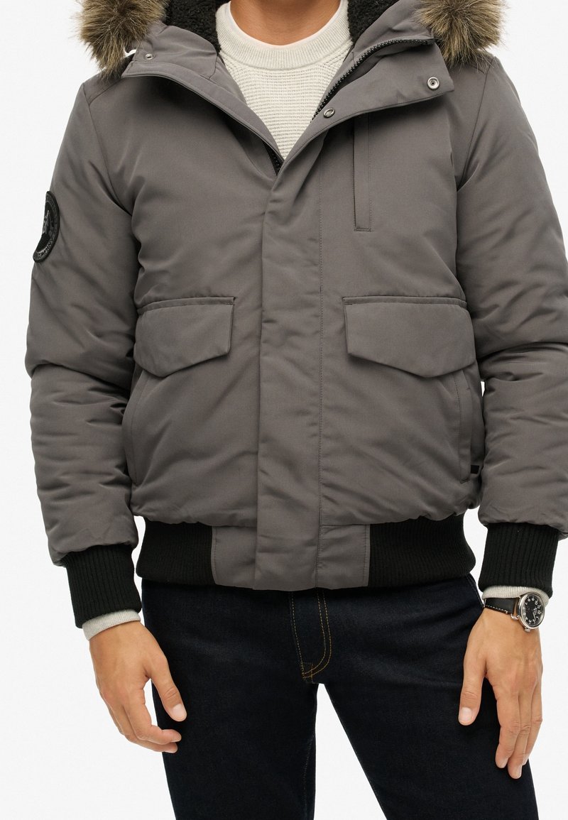 Graue gesteppte Jacke mit abnehmbarer, mit Fell gefütterter Kapuze, Druckknopfverschlüssen, zwei Fronttaschen, gerippten Bündchen und einem schwarzen Logopatch am Arm.