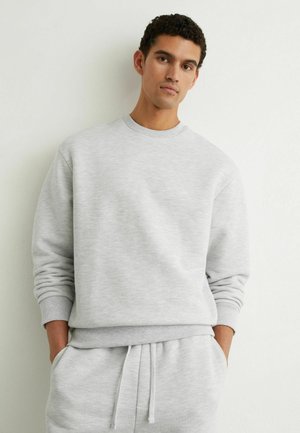 Młody mężczyzna z kręconymi włosami, ubrany w jasnoszarą bluzę i pasujące spodnie dresowe, z rękami w kieszeniach, stojący na tle jednolitej ściany.