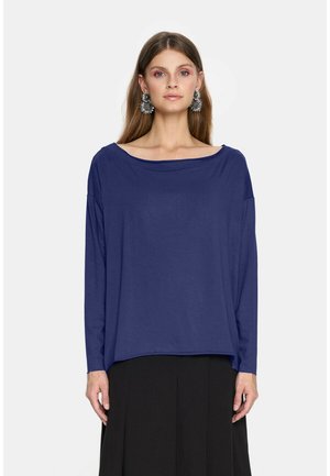 Kobieta ubrana w luźną granatową bluzkę z długim rękawem i czarną spódnicę, z dużymi srebrnymi kwiatowymi kolczykami, stoi na jednolitym tle.