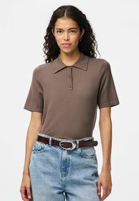Rjava polo majica s kratkimi rokavi z teksturirano površino in tremi gumbi na ovratniku. Kombinirana z modrimi kavbojkami z visokim pasom in rjavim pasom.
