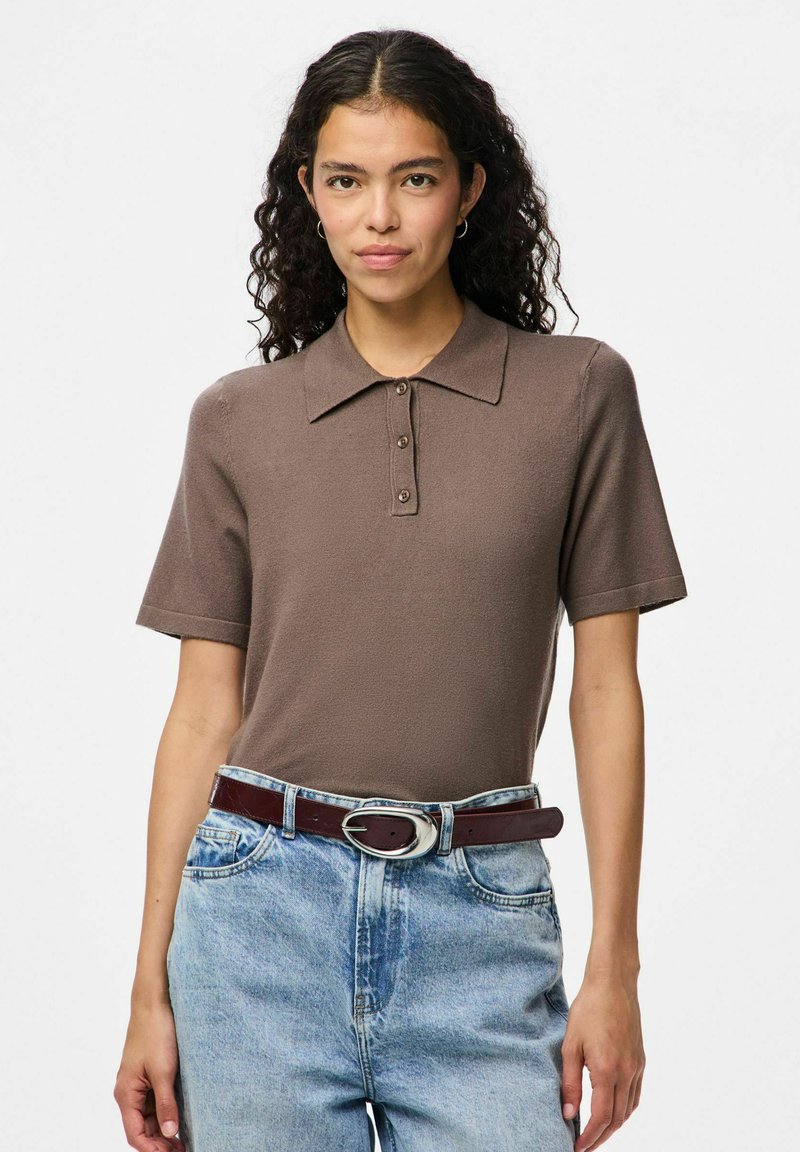 Rjava polo majica s kratkimi rokavi z teksturirano površino in tremi gumbi na ovratniku. Kombinirana z modrimi kavbojkami z visokim pasom in rjavim pasom.