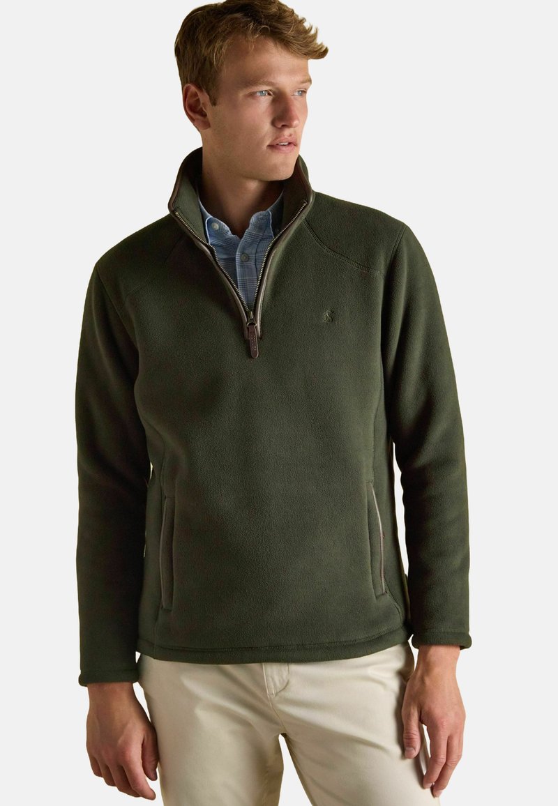 Uomo che indossa un pullover in pile verde oliva con zip a mezza lunghezza sopra una camicia a quadri blu chiaro e pantaloni beige, mentre guarda di lato.
