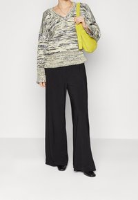 Person, der einen gemusterten Strickpullover, breite schwarze Hosen, schwarze Schuhe trägt und eine gelbe Umhängetasche vor einem einfarbigen Hintergrund hält.