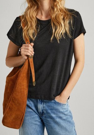 Frau mit welligem blondem Haar, die ein schwarzes T-Shirt und blaue Jeans trägt und mit der linken Hand den Riemen einer beigen Wildleder-Schultertasche hält.