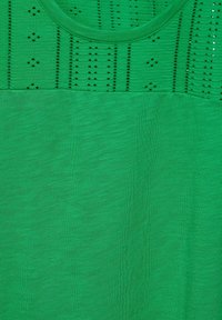Maglietta verde con scollatura rotonda. La parte superiore presenta una texture a motivo con ritagli; la parte inferiore è in tessuto verde liscio e uniforme.
