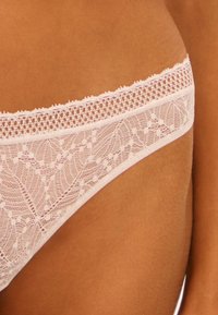 String en dentelle rose doux avec un motif floral et une taille festonnée, présentant des détails texturés complexes et des bords lisses.