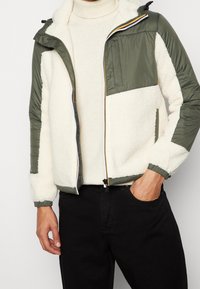 Sherpa-gefütterte Jacke mit olivgrünem Außenmaterial, Reißverschluss, Taschen und elastischen Bündchen für eine körpernahe Passform.