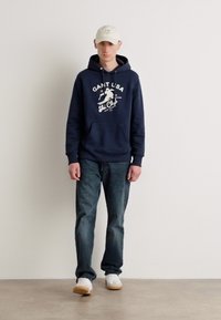 Sudadera navy con logo blanco del GANT USA Ski Club, bolsillo frontal, combinada con jeans oscuros y una gorra beige. El calzado son zapatillas blancas con suelas de goma.