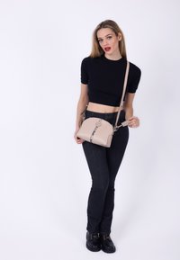 Ung kvinde i sort crop top og jeans holder en beige crossover-taske med slange-mønsterstribe mod hvid baggrund.