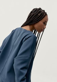 Ciemnoniebieska bluza z gładkiego materiału, z opadającymi ramionami i widocznymi detalami szwów. Długie warkocze częściowo widoczne.