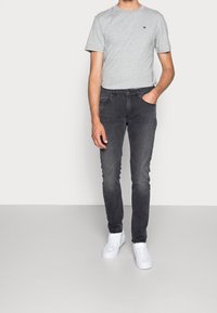 Camiseta gris de manga corta con un pequeño logo, combinada con jeans ajustados gris oscuro. Zapatillas blancas completan el conjunto.