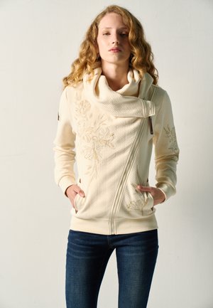 Mujer con chaqueta bordada color crema con cuello alto doblado y vaqueros azul oscuro, de pie con las manos en los bolsillos frente a un fondo liso.