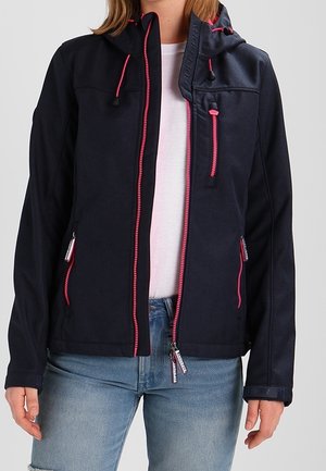 Zwarte softshell jas met roze ritsen, drie met rits afsluitbare zakken, gedragen over een wit overhemd en blauwe spijkerbroek, bij een staande persoon.