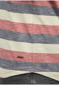 Stribet T-shirt med røde, cremefarvede og marineblå vandrette striber, der har et lille sort mærke med røde accenter nederst til venstre.