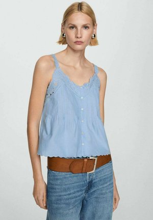 Eine Frau trägt ein hellblaues ärmelloses Oberteil mit Knöpfen und Spitzendetails, kombiniert mit blauen Jeans und einem breiten braunen Gürtel.