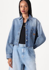 Giacca di jeans cropped di blu medio con chiusura a bottoni, taschino sul petto e leggera usura. Indossata sopra una maglietta nera e jeans blu chiaro.