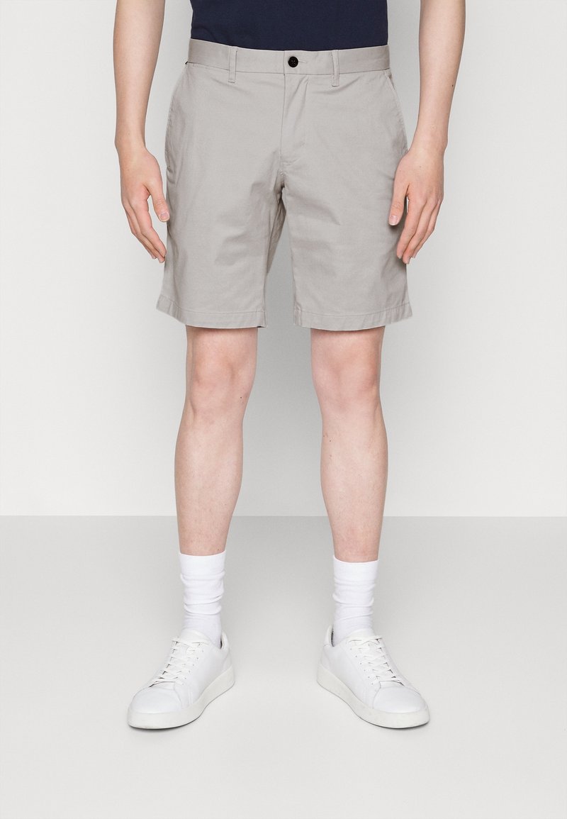 Pantaloni corti di cotone beige con fronte piatto, tasche laterali e chiusura con bottone. Abbinati a sneakers bianche e calzini bianchi alla caviglia.
