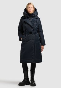 Temno modra podložena trenč plašč z velikim kapuco, dvorednimi gumbi, pasom in elegantno, gladko teksturo. Stiliran s črnimi legicami in škornji.