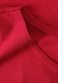 Tissu rouge avec une texture lisse ; présente un col rond et un bord replié. La surface montre de légères rides et des détails de couture.