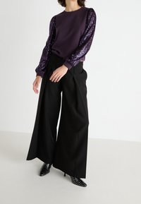Sudadera morada con mangas de lentejuelas, combinada con pantalones negros de pierna ancha. Zapatos de tacón negro de punta con lunares blancos completan el conjunto.